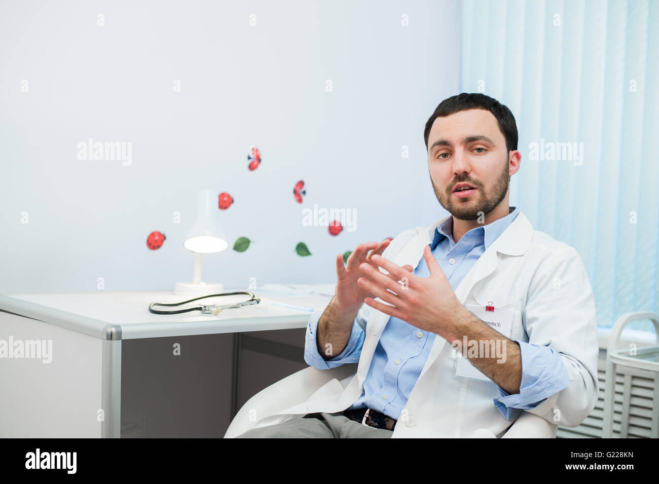 Cheerful doctor talking and looking into the camera. Physician in conversation with patient while sitting at hospital. Stock Photo