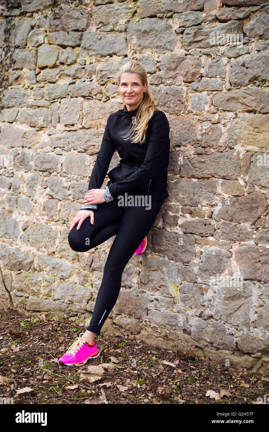 A middle aged female runner posing by leaning against a stone wall. Stock Photo