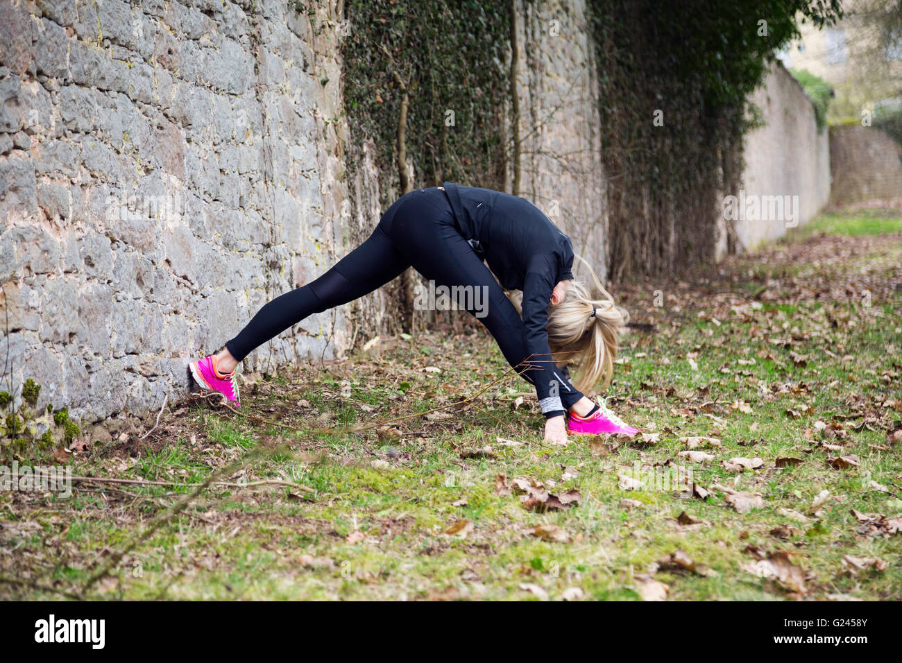 Middle aged woman stretching in the park against a grey wall before a run Stock Photo