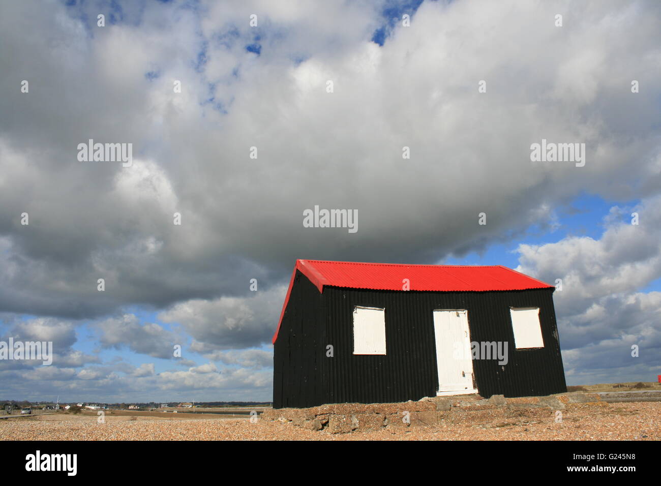 A LANDSCAPE VIEW AT RYE HARBOUR NATURE RESERVE OF A RED ROOFED FISHEMANS HUT BY THE RIVER IN THE SUNSHINE WITH INTERESTING CLOUD Stock Photo