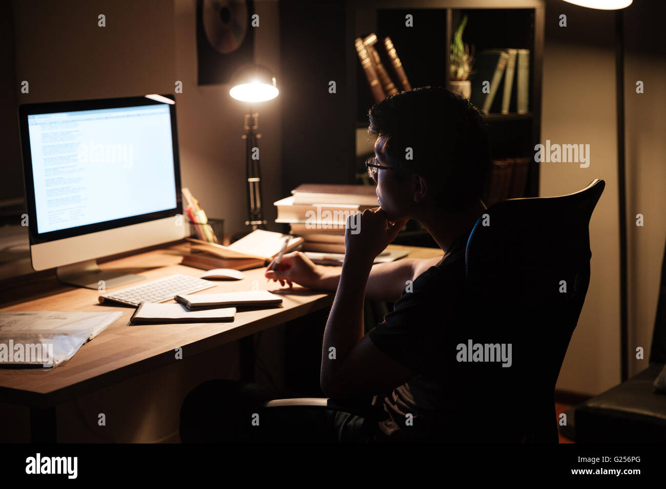 Thoughtful asian young man sitting and studying using computer in dark room at home Stock Photo