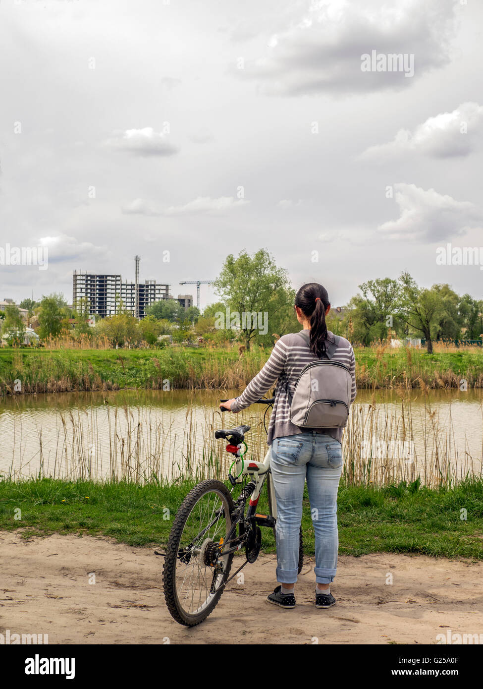 Woman standing by lake with bicycle looking at new building construction Stock Photo