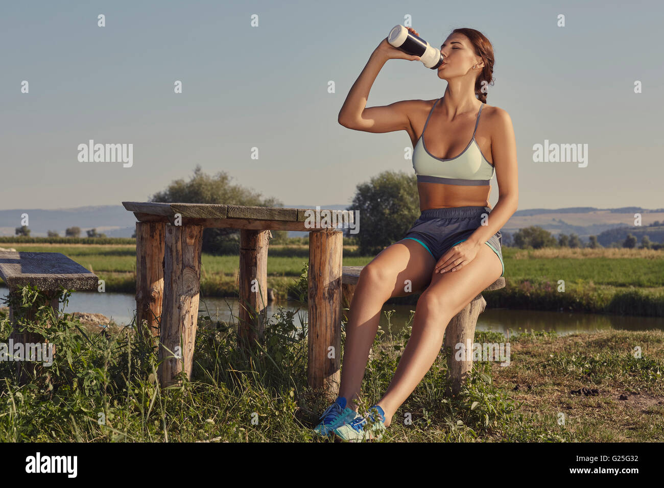 Attractive young lady in summer sportswear drinks water or energizing shake after the morning run while resting on a rustic wood Stock Photo
