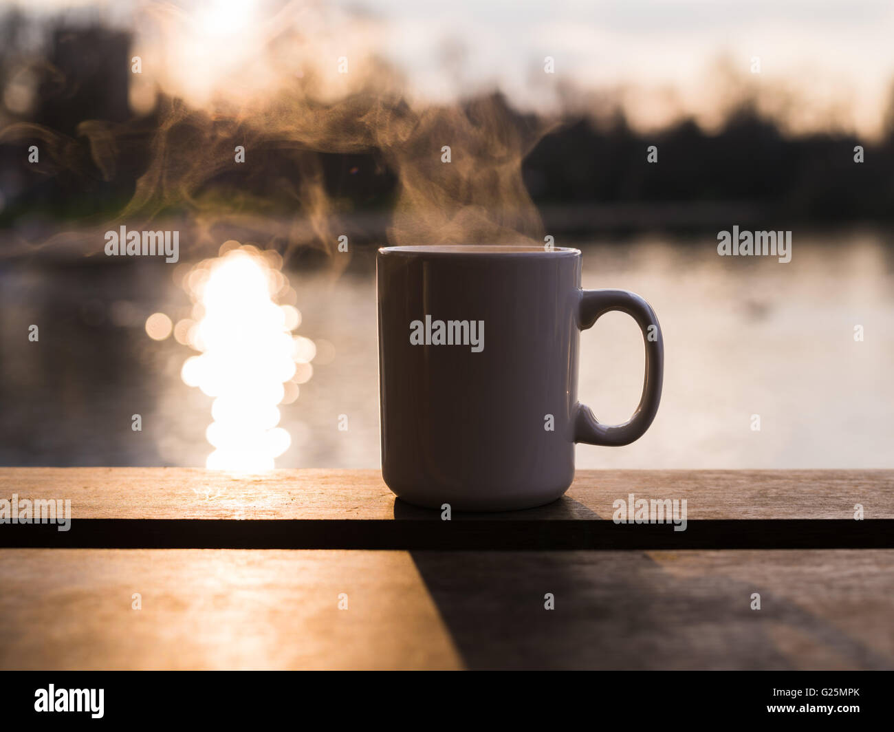 Steam rising from a mug with a hot beverage by a pond at sunset in the ...
