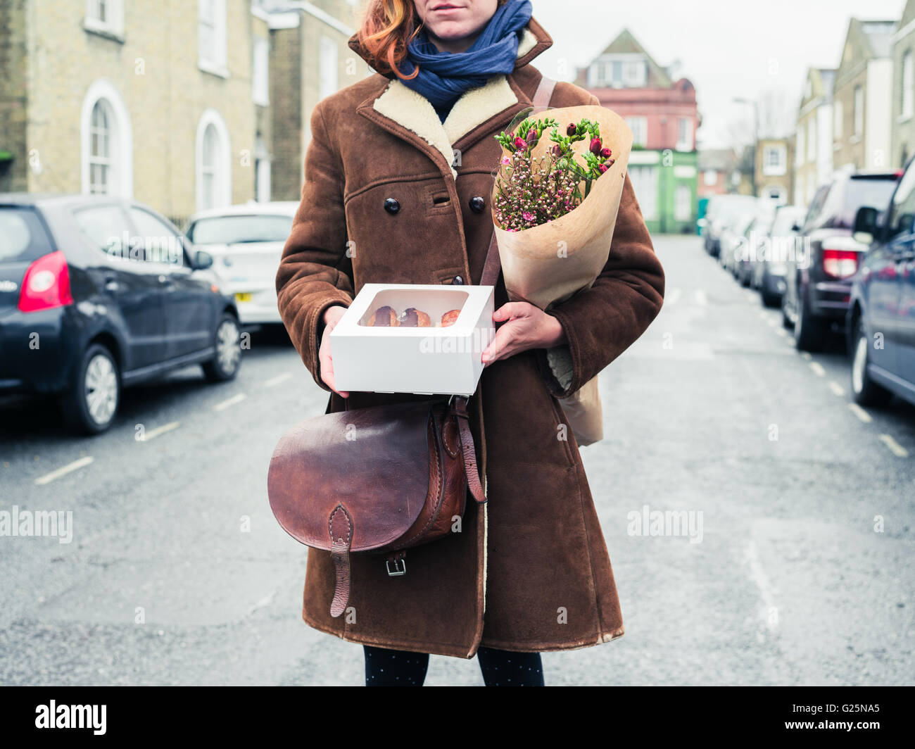 A young woman is standing in the street with a box of cake and a bouquet of flowers Stock Photo