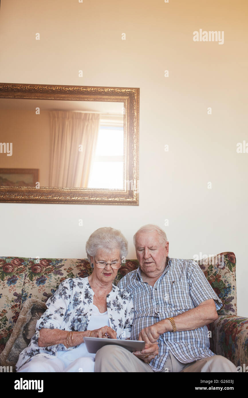 Vertical indoor shot of retired couple at home using digital tablet. Senior caucasian man and woman sitting together on sofa at Stock Photo