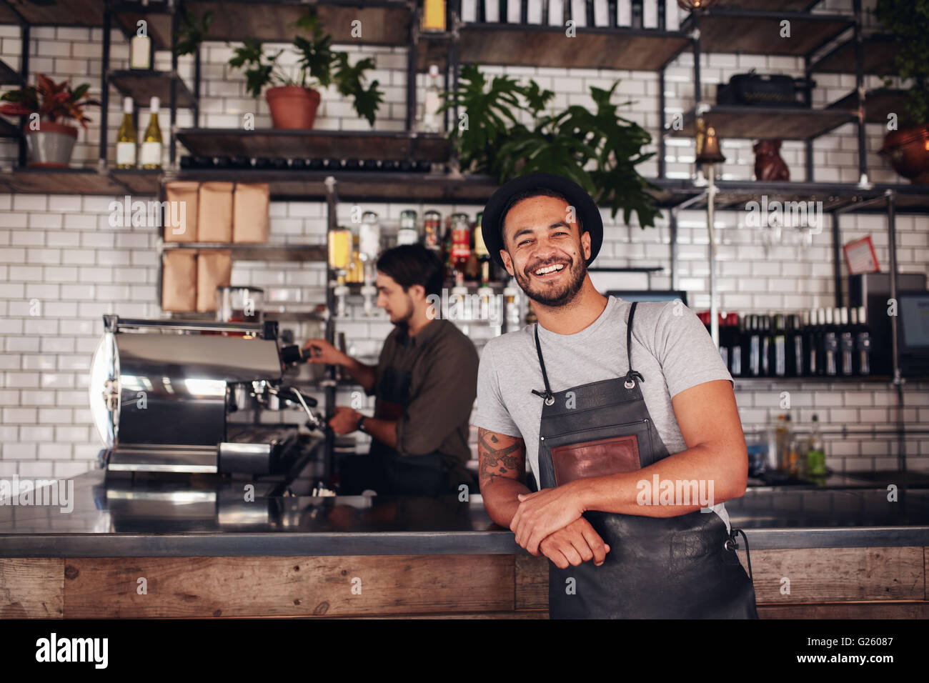 Coffee shop worker smiling to camera, standing at the counter. Happy young man in apron and hat leaning to cafe counter, with wa Stock Photo