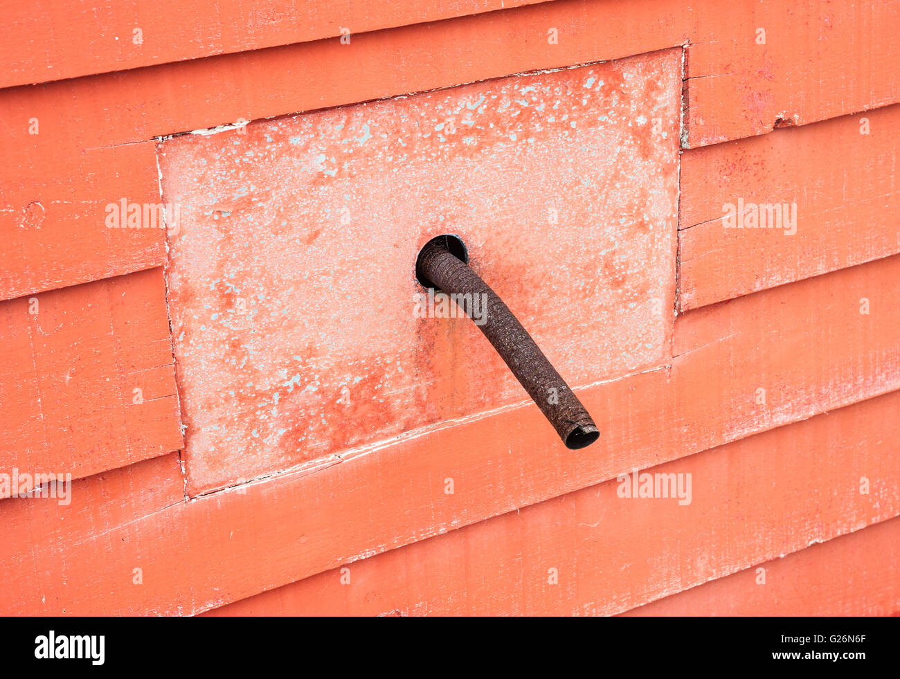 Rusted old curved metal pipe protruding from square panel in red wall. Stock Photo