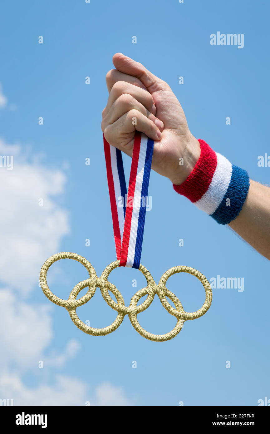 RIO DE JANEIRO - FEBRUARY 4, 2016: Hand with red white and blue wristband holding gold Olympic rings medal hanging in blue sky. Stock Photo