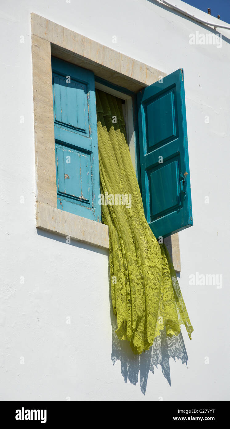 a lime green net curtain flowing out of an open window in a square frame  with blue shutters in a white washed rendered wall, Greece Stock Photo