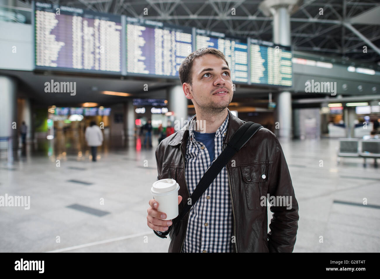 Smiling thoughtful man in front of arrivals and departures board at the airport with a coffee Stock Photo