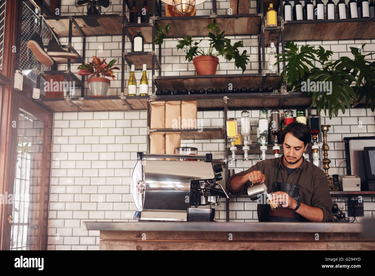 Indoor shot of young male barista making a cup of coffee while standing behind cafe counter. Young man pouring milk into a cup o Stock Photo