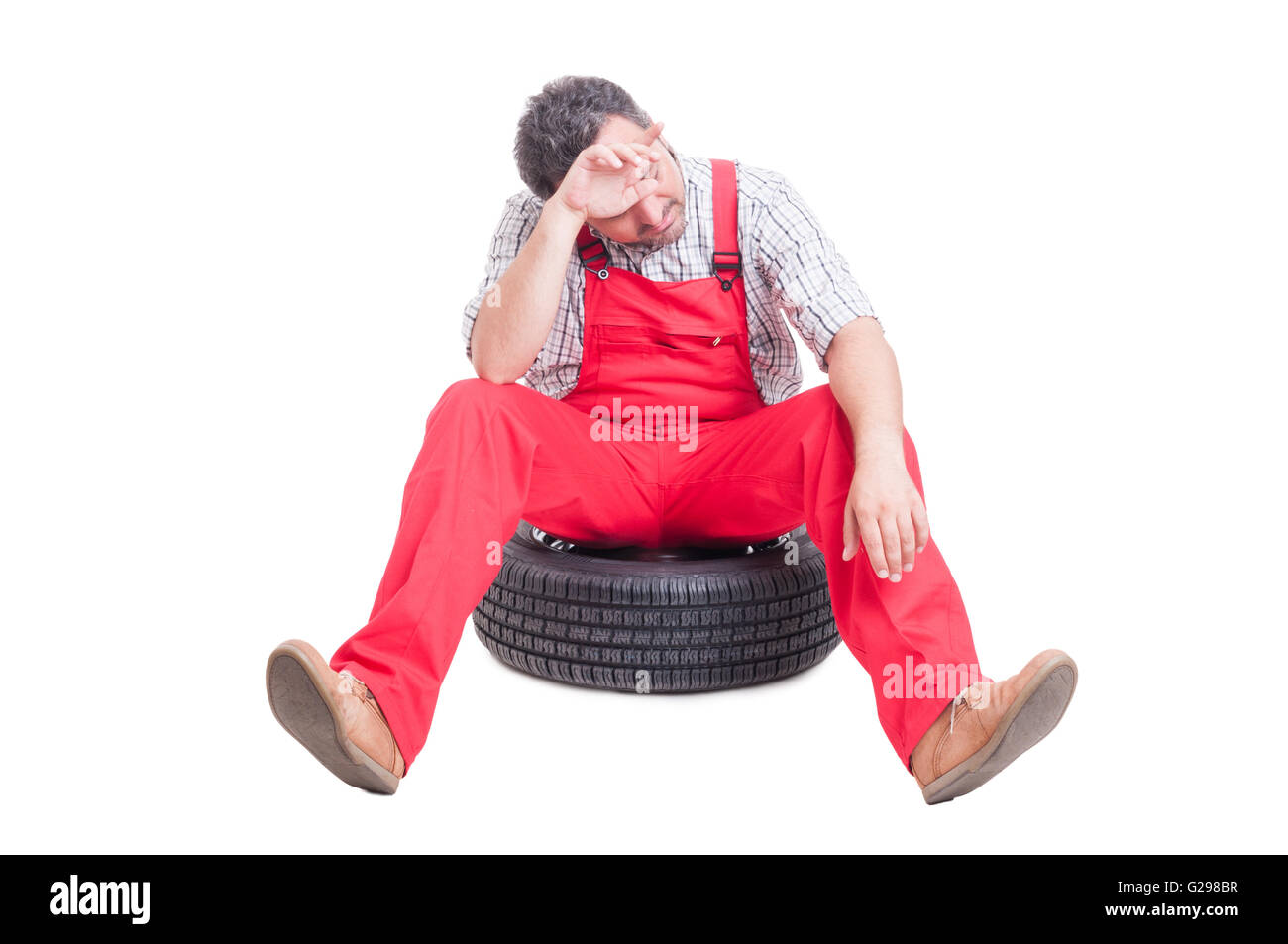 Tired and exhausted mechanic sitting on a car wheel wiping his sweaty forehead Stock Photo