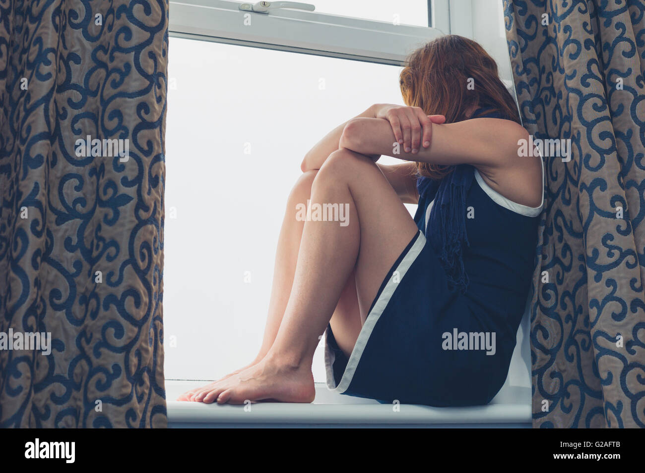 A young woman is sitting on a window sill and is looking out on a rainy day Stock Photo