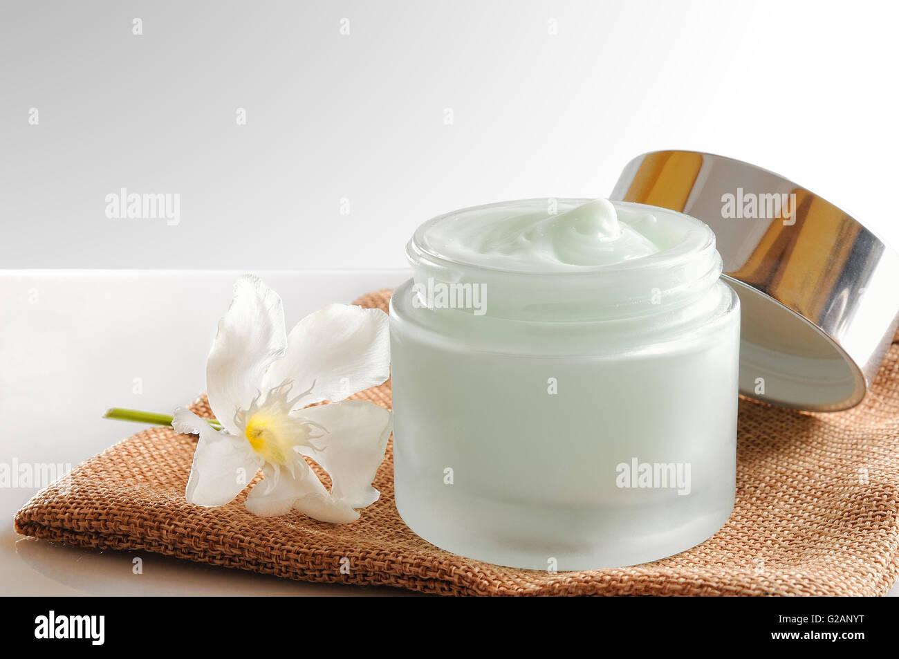 Glass open jar with facial or body cream on burlap. With lid and flower.Isolated background. Front view. Stock Photo
