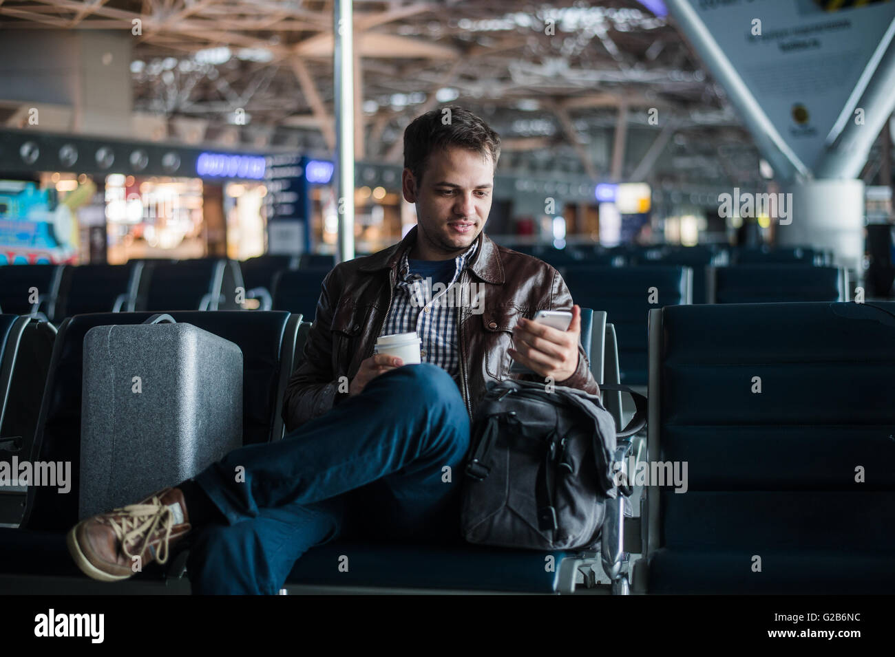 Handsome smiling man in casual wear holding luggage and messaging through his mobile phone while sitting at the hall of airport with coffee cup Stock Photo