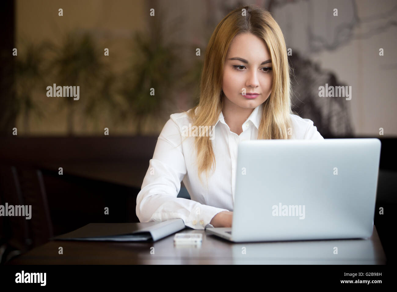 Young beautiful blond caucasian businesswoman working on laptop, typing with focused expression, cellphone and black folder Stock Photo