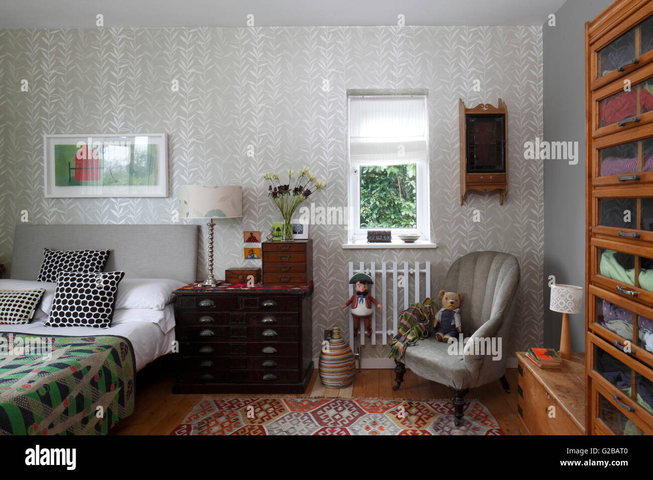 Bedroom with silver patterned wallpaper and a vintage shop fitting clothes storage. Black and white pillows on bed and grey chair in corner of room with stuffed animals. Stock Photo
