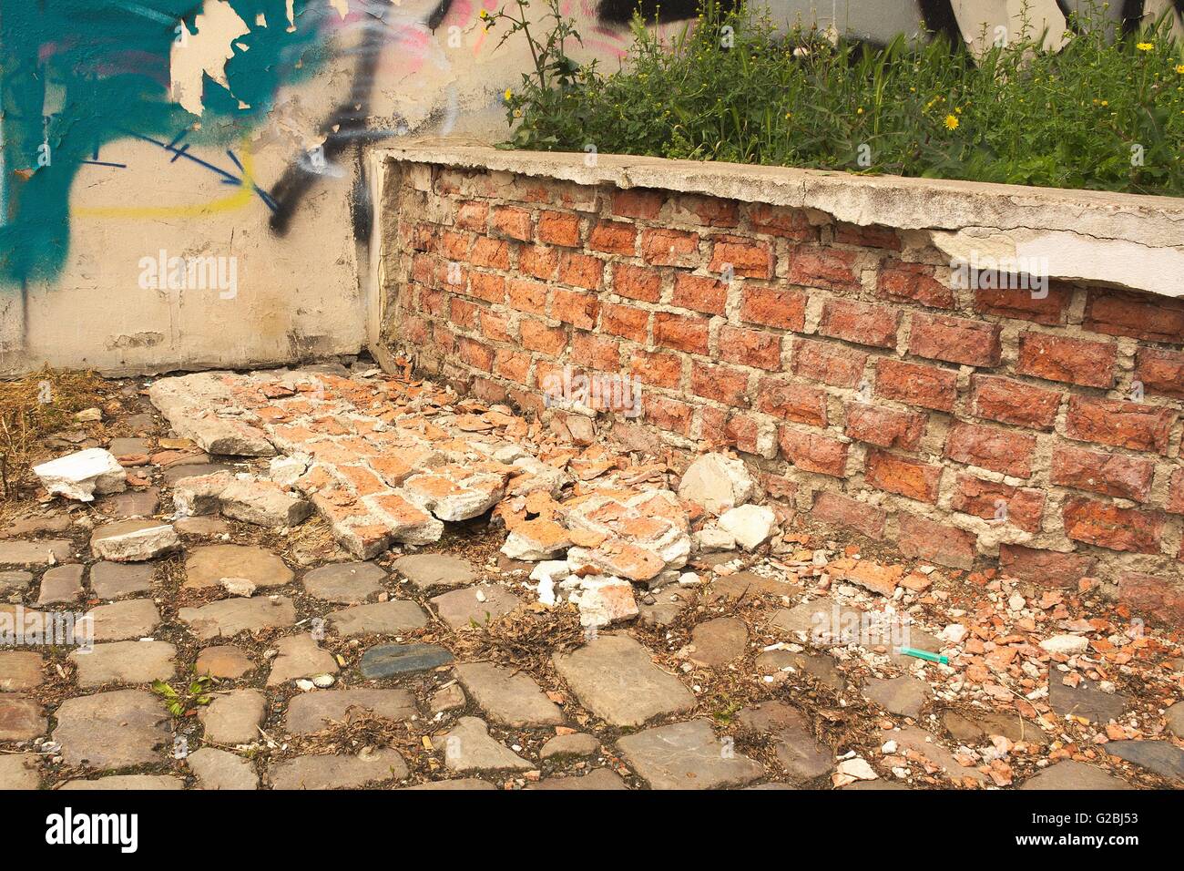 Low disintegration of the old brick wall. Brick lying on a paved street.On the wall is visible graffiti Stock Photo