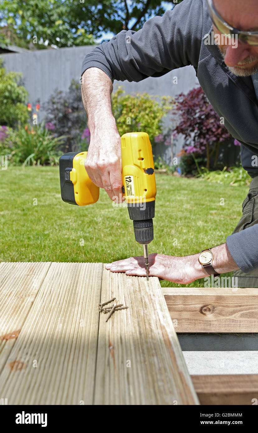 A workman uses a cordless drill to assemble garden decking in an English garden featuring a lawn and shrubs in the background Stock Photo