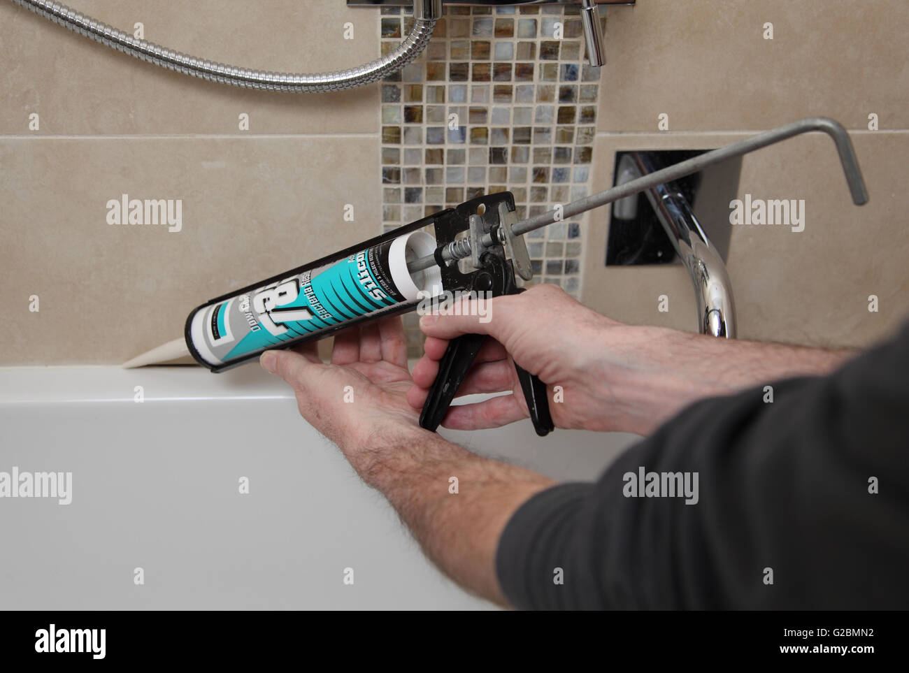 A plumber uses a mastic gun to apply stone-coloured silicone sealant between tiles and bath edge in a modern, tiled bathroom Stock Photo