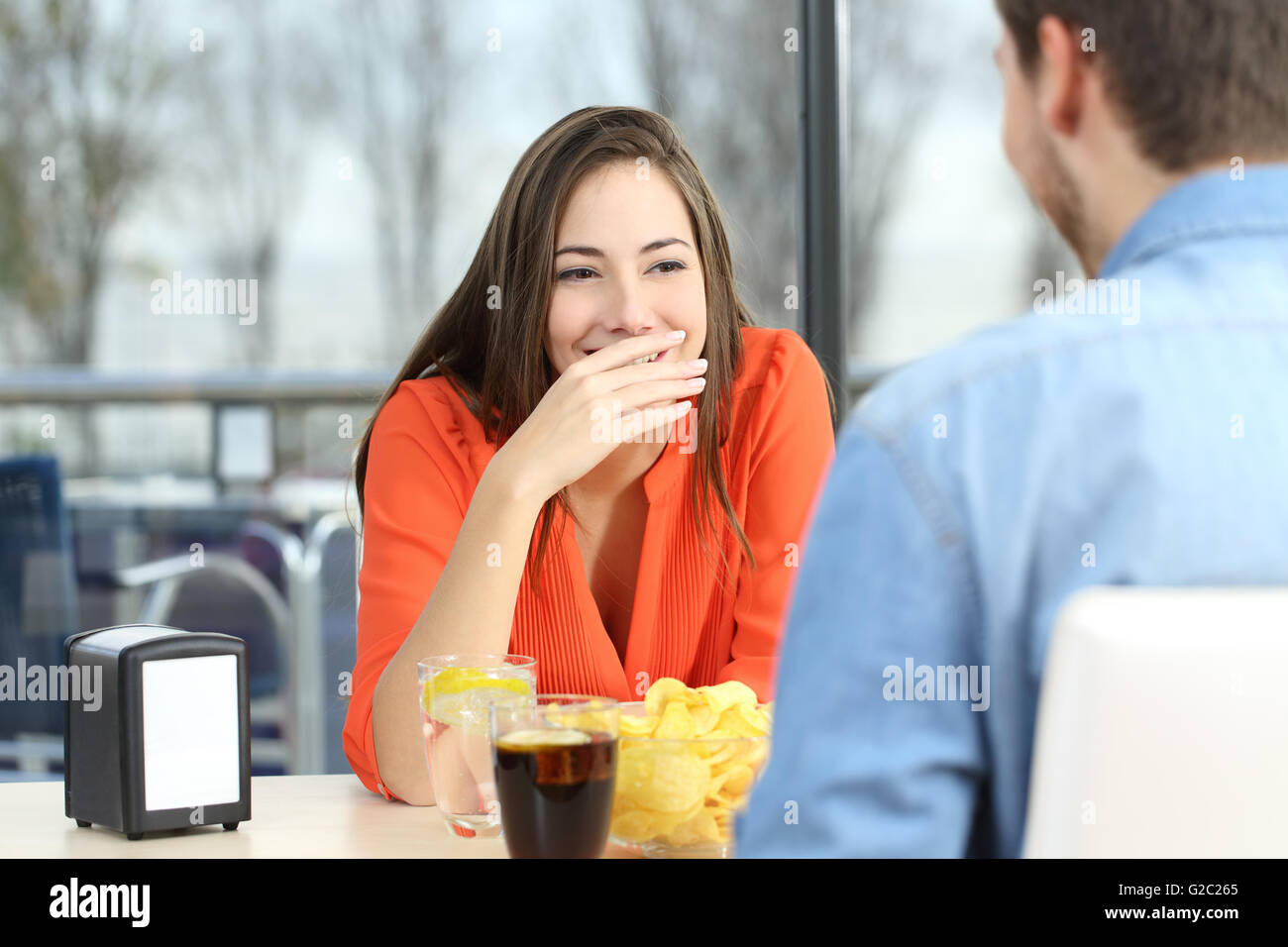 Woman covering her mouth to hide smile or bad breath during a date in a coffee shop with a window in the background Stock Photo
