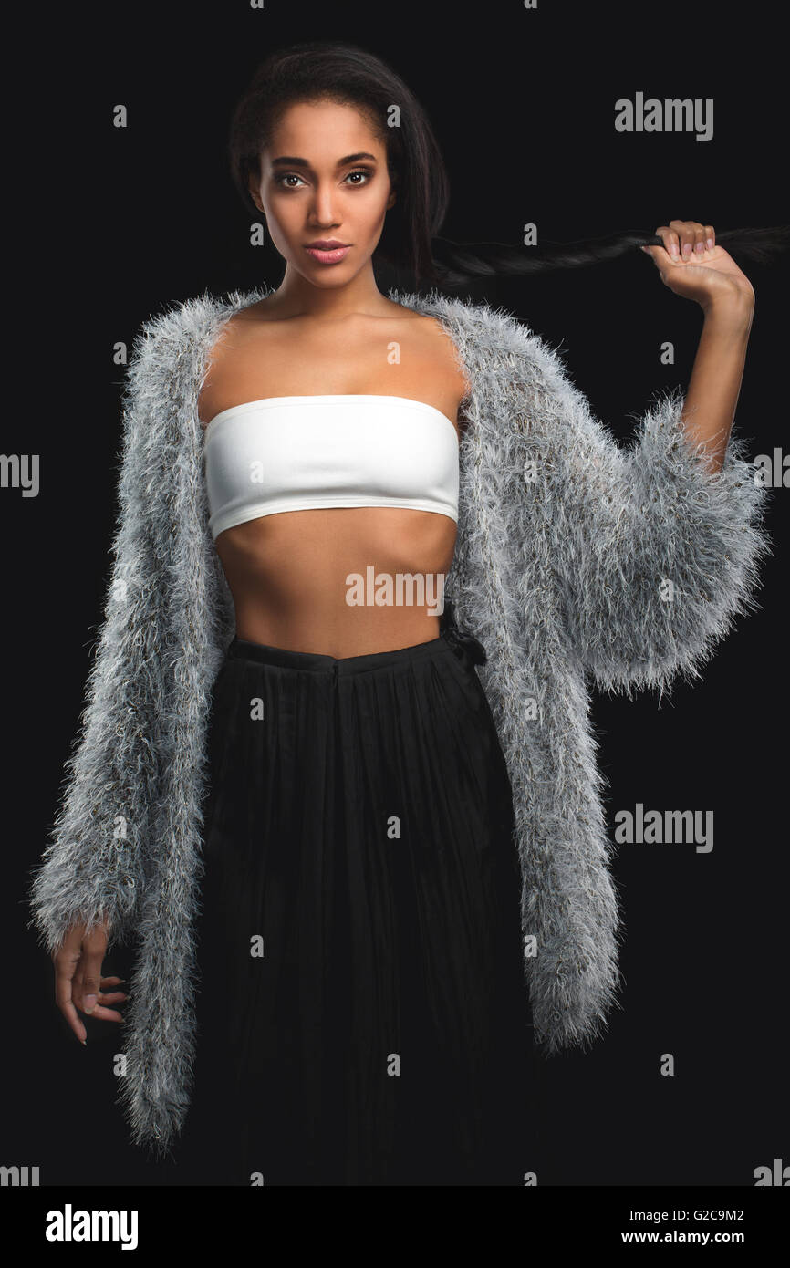 Fashionable mulatto in fur coat holding her braid hairstyle on black background. African Amarican girl with long hair looking at camera. Stock Photo