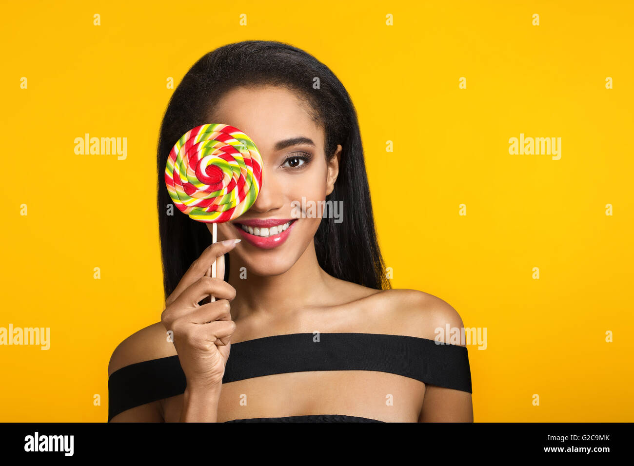 Smiling cute mulatto girl holding and covering her eye with big colourful sweet lollipop looking at camera over sunny, yellow background. Stock Photo