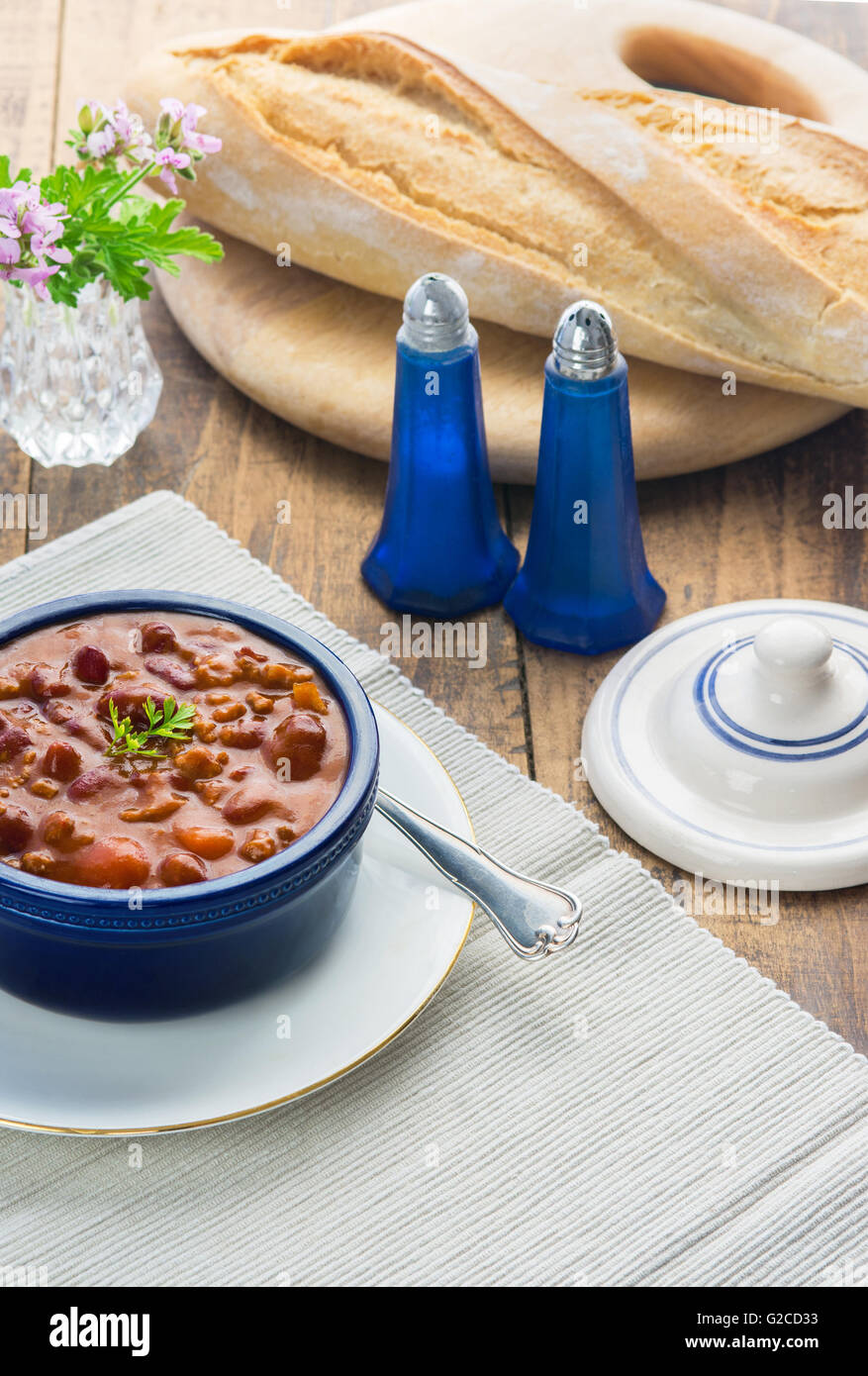 vertical image of a table setting showing a meal of chilli con cane on a wooden table set with bread in a bright window light. Stock Photo