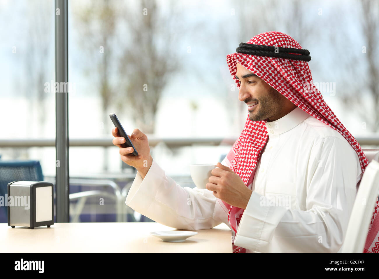 Side view of an arab man texting in a smart phone in a coffee shop with a window with a sunny day in the background Stock Photo