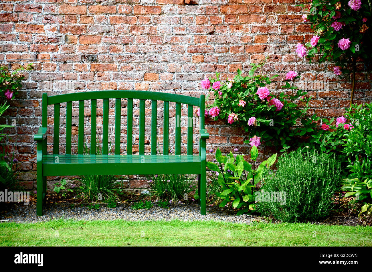 Green Park Bench Against Brick Wall Surrounded by Pink Roses and Plants Stock Photo