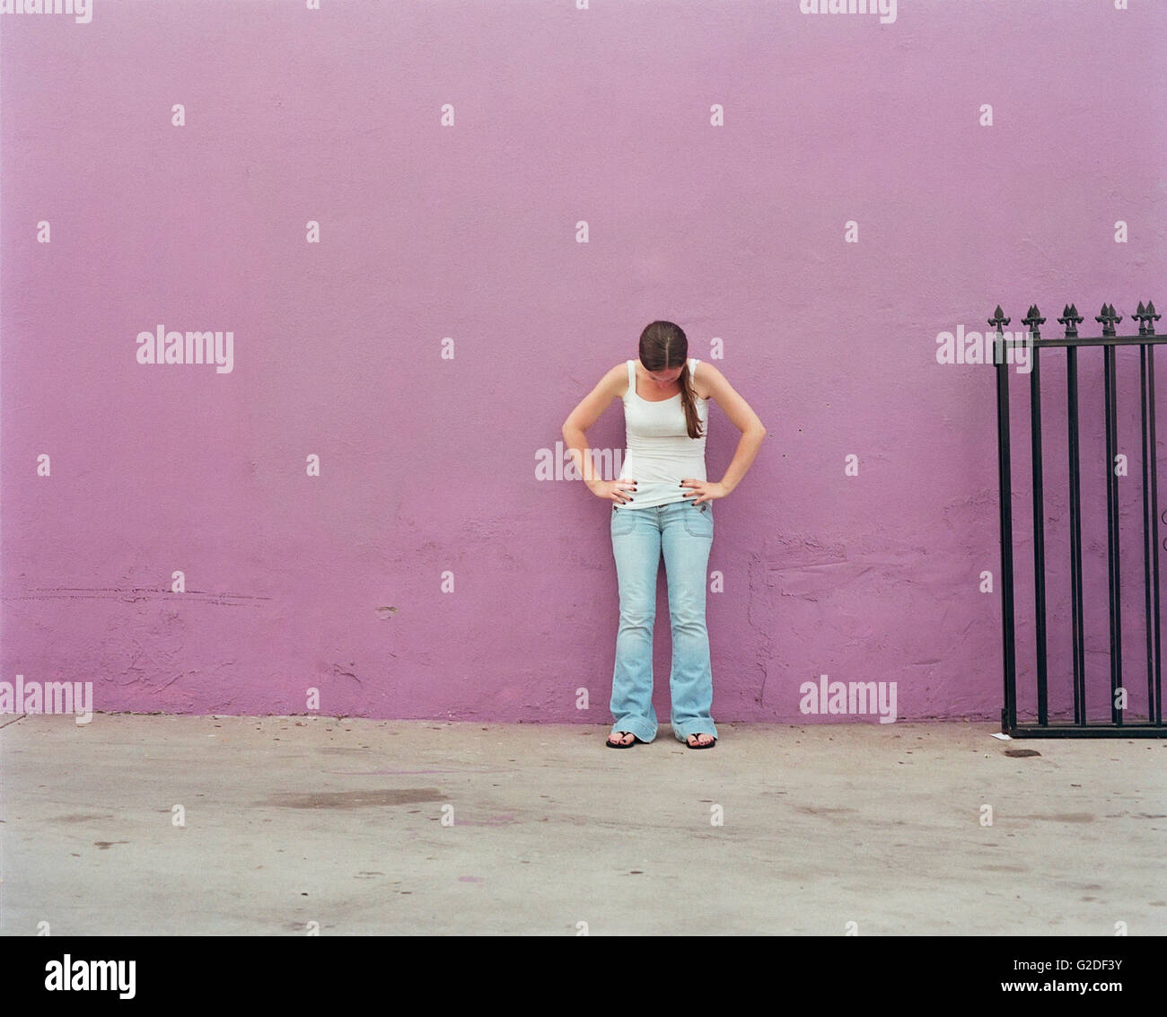 Woman in Tank top and Jeans Standing Against Purple Wall with Hands on Hips and Looking Down Stock Photo