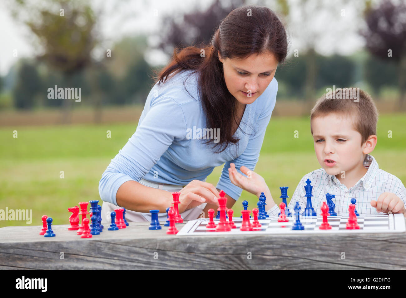 Elegant young boy in white shirt and his mother learning to play chess with blue and red chess pieces on wood table in the park Stock Photo