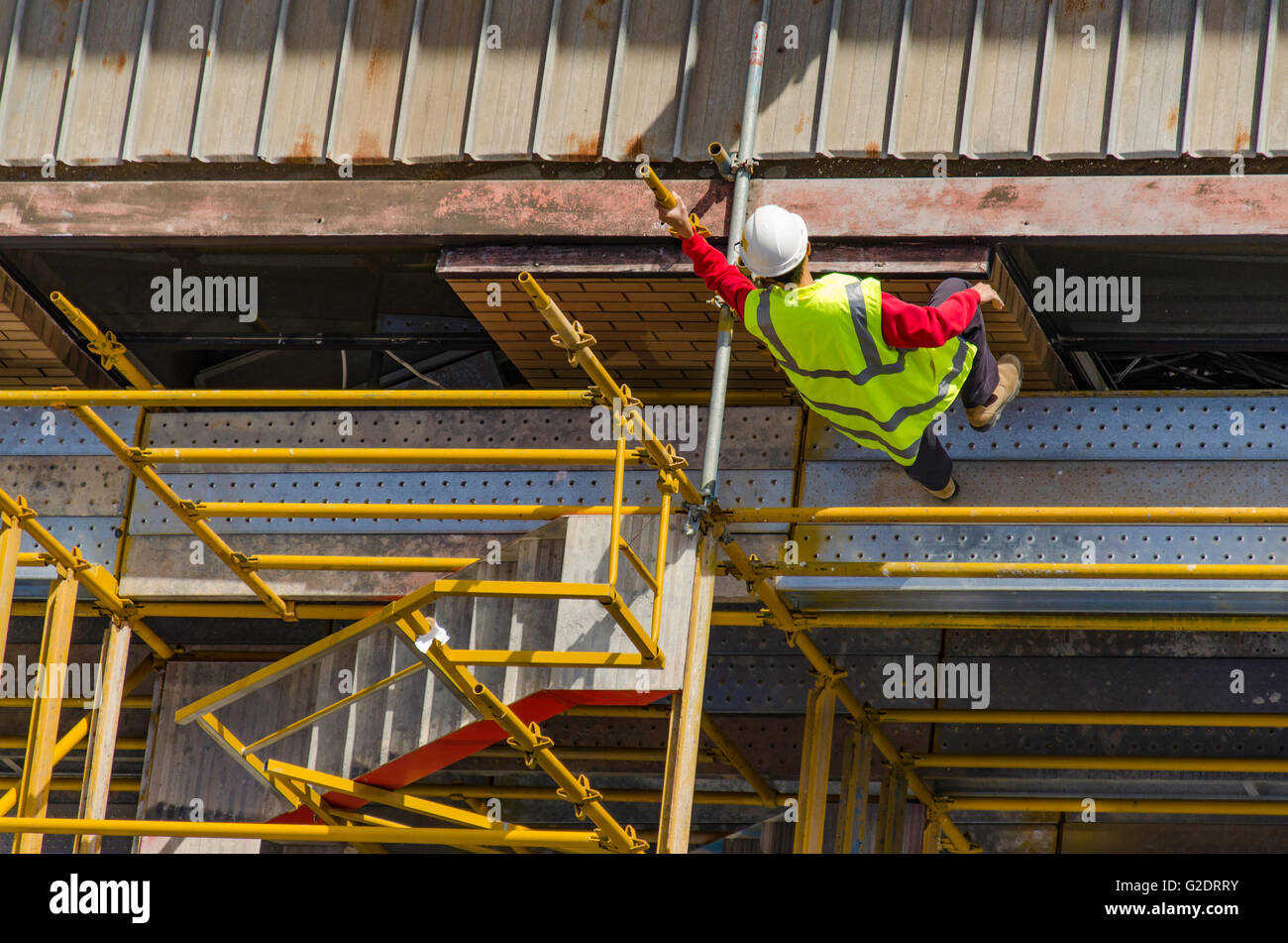 A white male worker in high visibility (hi-vis) apparel climbing on the steel roof of a multi-story building site, on yellow coloured scaffolding Stock Photo