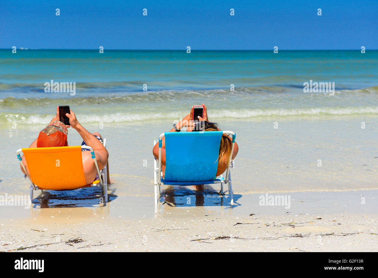 Preoccupied couple can't stop looking at their cell phones to enjoy each other or the beach shoreline while reclining in chairs Stock Photo
