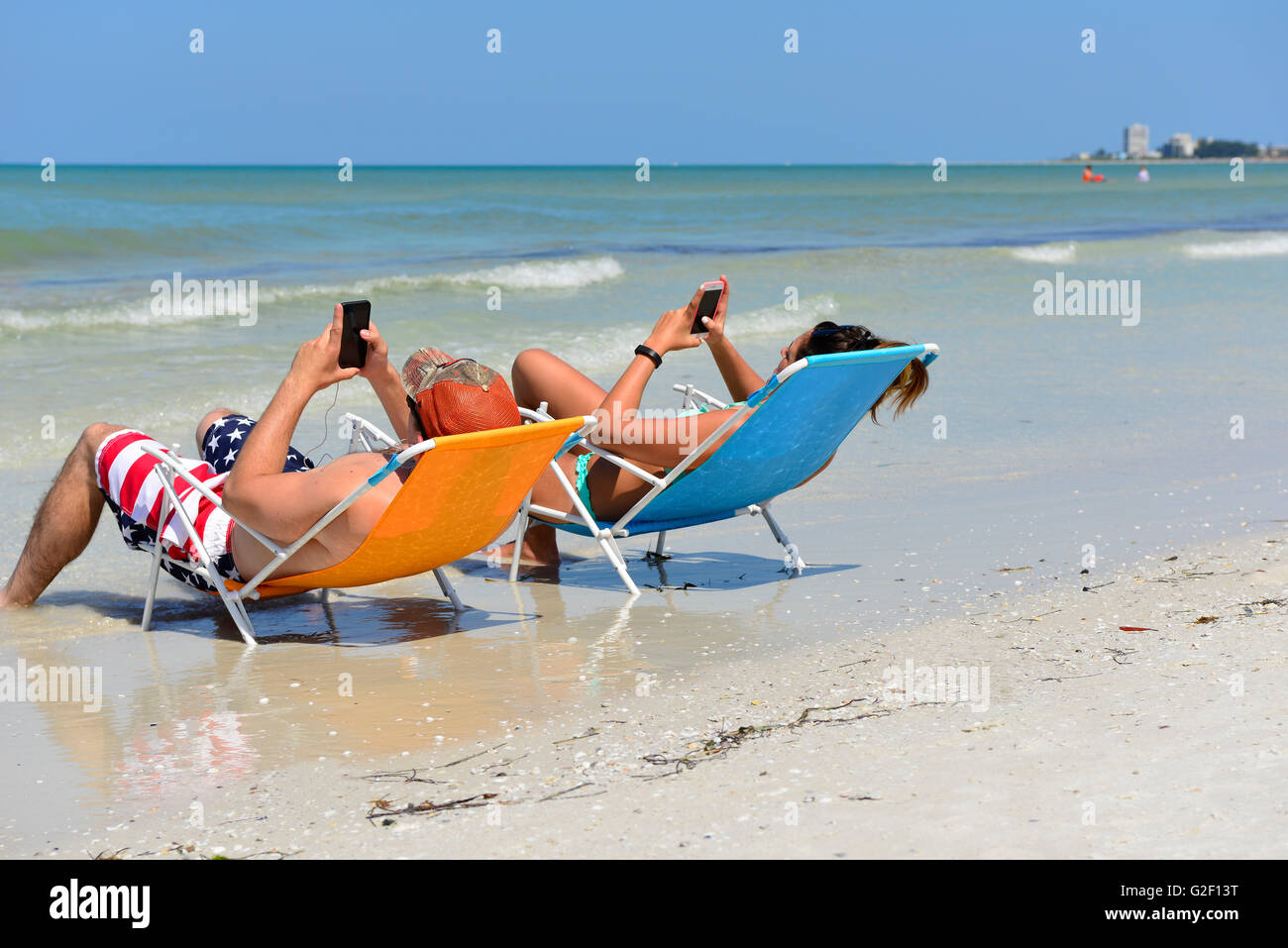 Preoccupied couple can't stop looking at their cell phones to enjoy each other or the beach shoreline while reclining in chairs Stock Photo