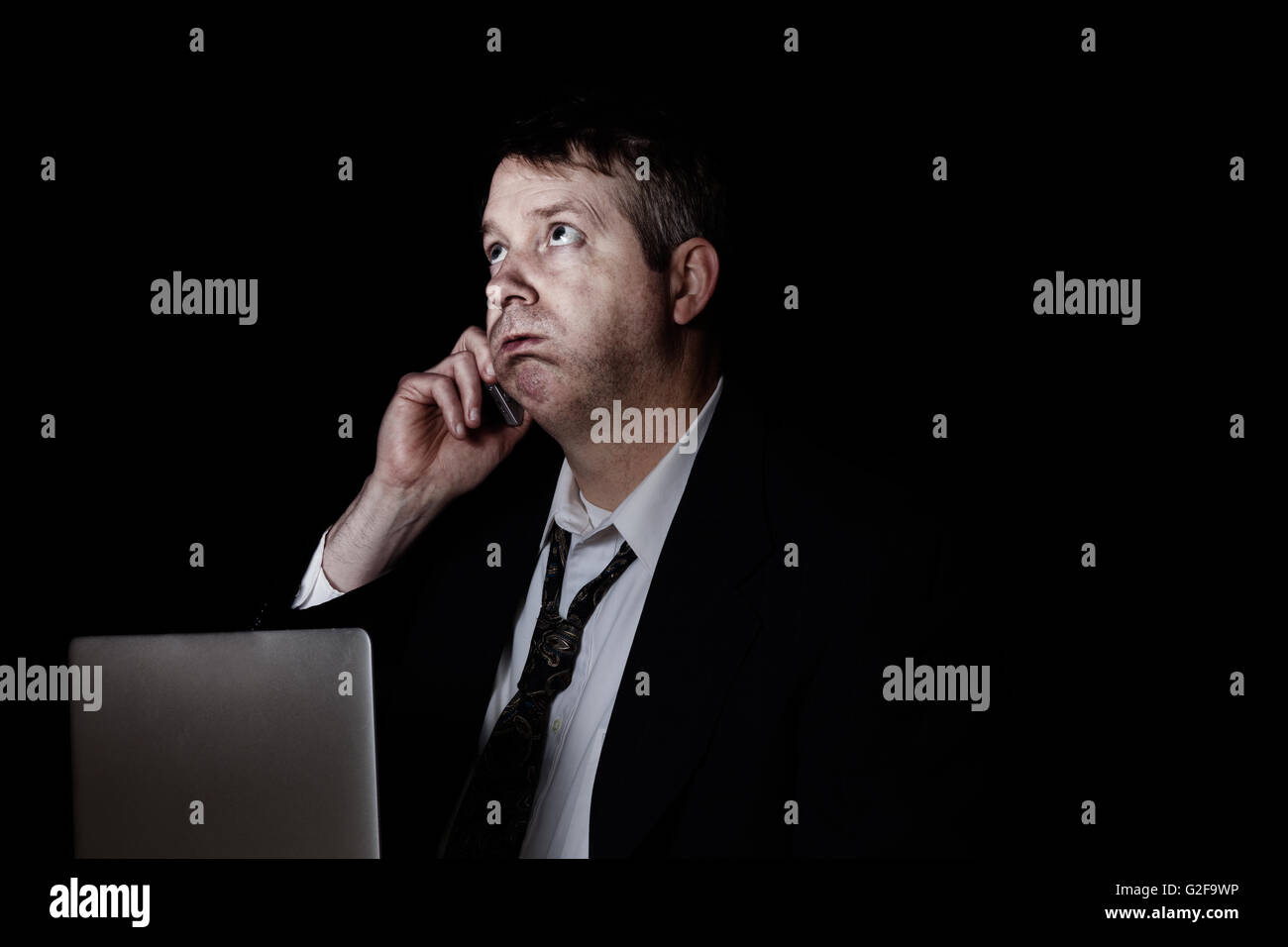 Side view of stressed business man on cell phone while working.  Dark background with light on face. Stock Photo