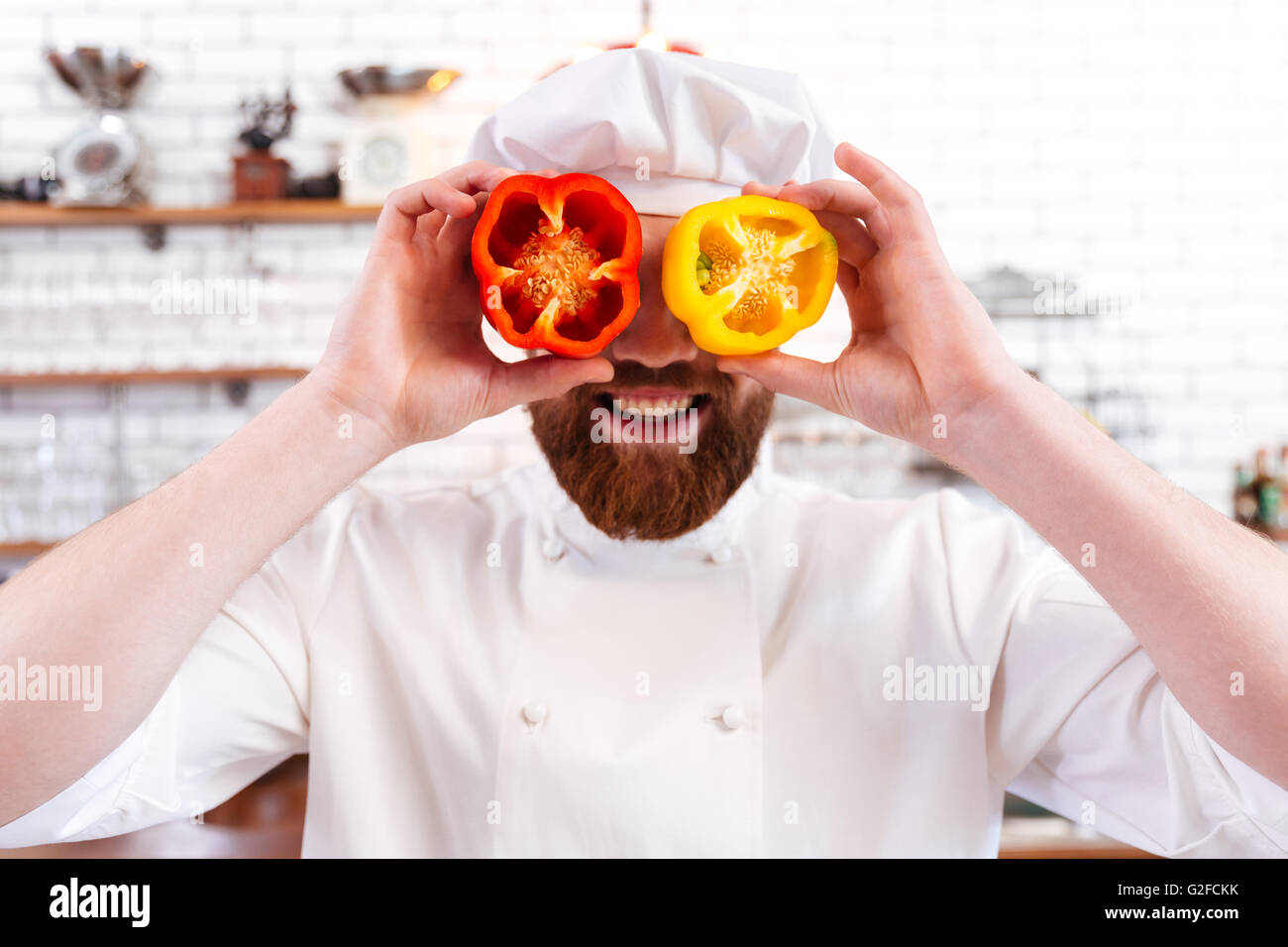 Playful bearded chef cook holding halves of red and yellow bell peppers in front of his eyes and smiling Stock Photo