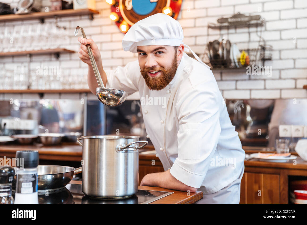 Cheerful bearded chef cook cooking and tasting soup from pan using ladle on the kitchen Stock Photo