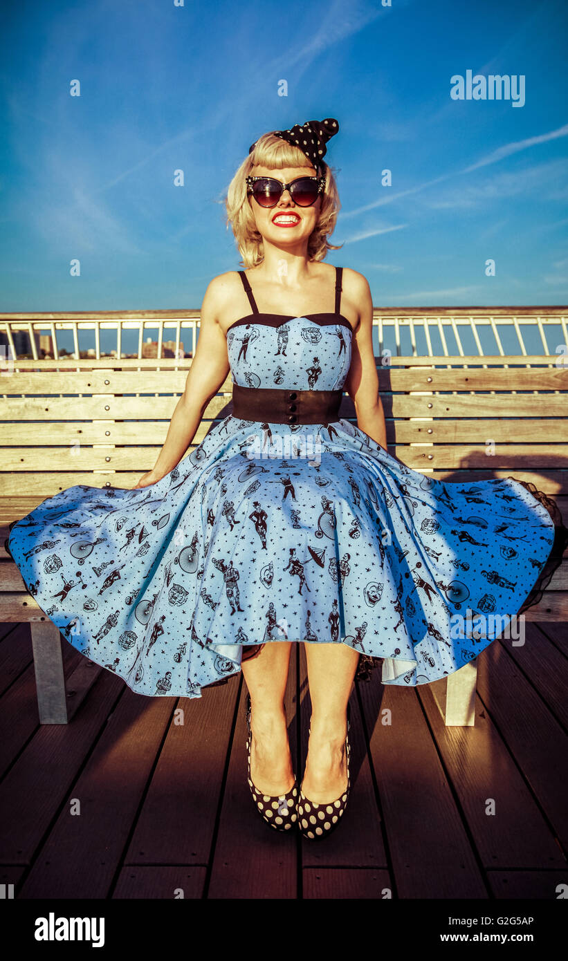 Young Adult Woman in Retro Dress and High Heels Sitting on Boardwalk Bench at Beach Stock Photo