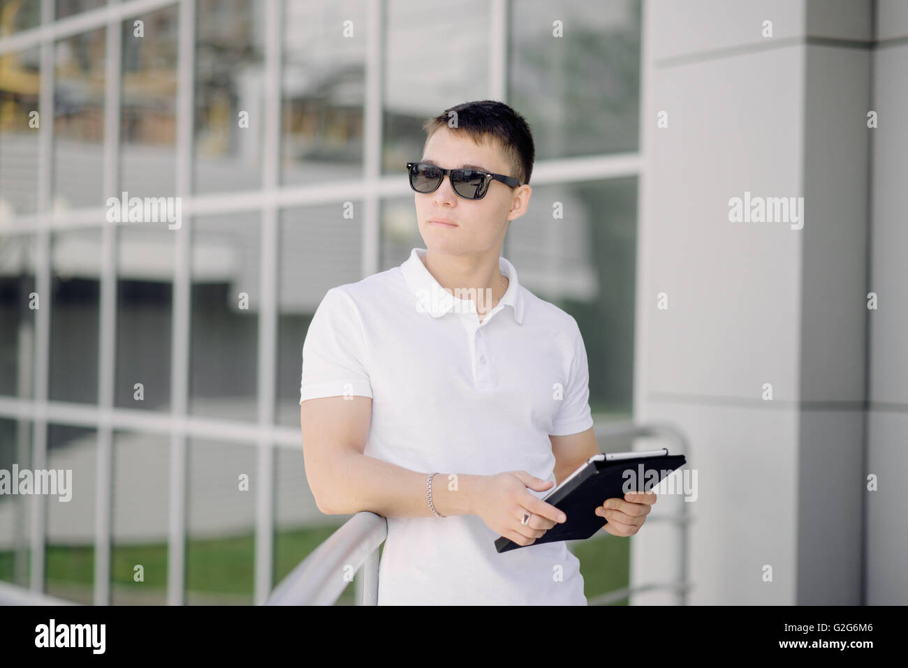 Portrait of a young businessman keeping tablet pc on the street. Man with dark sunglasses and white polo. Stock Photo