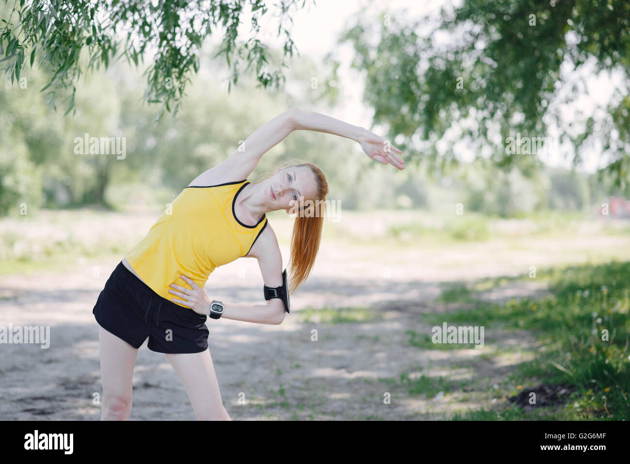 Young fitness woman runner stretching body before run. Sportsman standing under tree. Stock Photo
