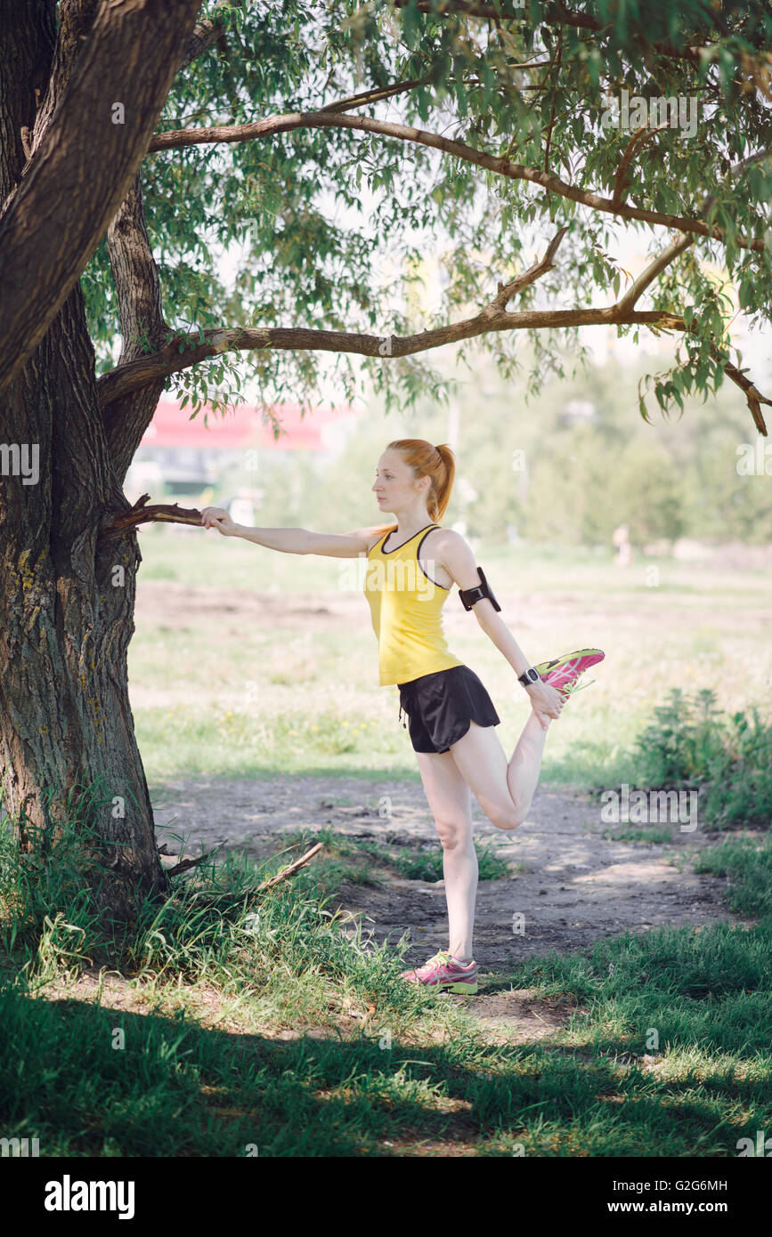 Young fitness woman runner stretching legs before run. Sportsman standing under tree. Stock Photo