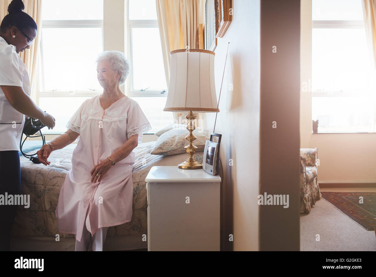 Indoors shot of senior woman's blood pressure being measured by nurse in nursing home. Home caregiver testing blood pressure of Stock Photo