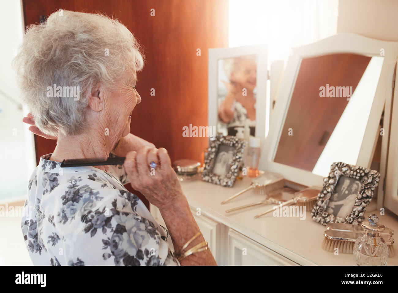 Happy senior woman combing her hair at dresser in house. Elderly female getting ready in front of a mirror at home. Stock Photo