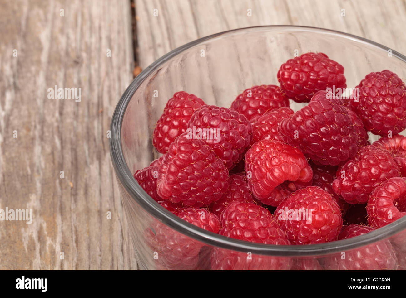 Clear glass bowl of ripe raspberries on a rustic farm picnic table in summer. Stock Photo