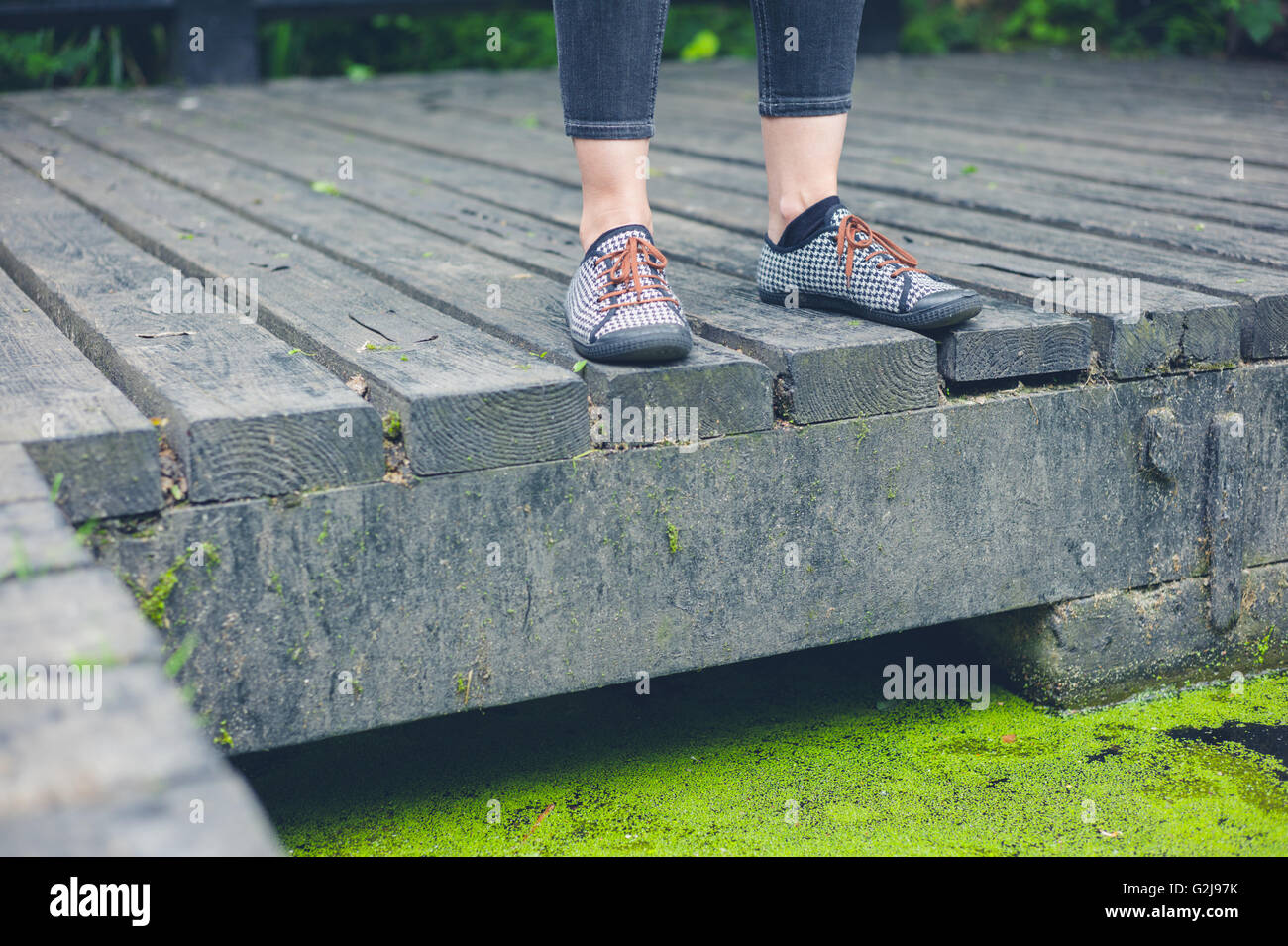 The feet of a young woman standing on a wooden deck by a dirty pond with algae Stock Photo