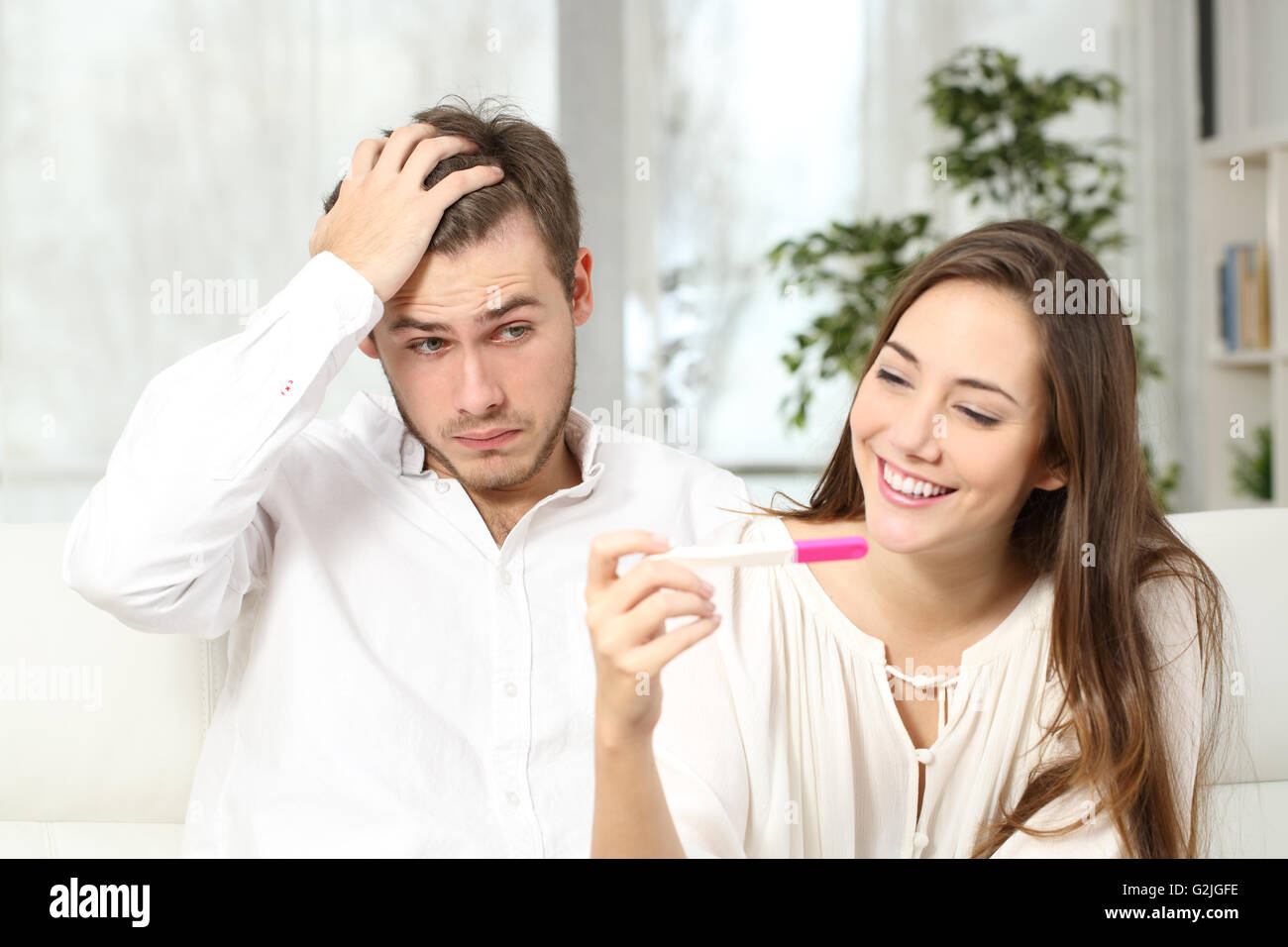 Not ready worried man checking a pregnancy test with his excited wife sitting on a couch at home Stock Photo
