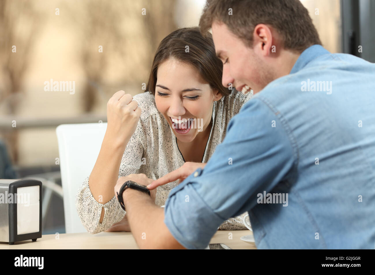 Excited couple reading e-mail with good news in a smartwatch in a coffe shop with a window in the background Stock Photo