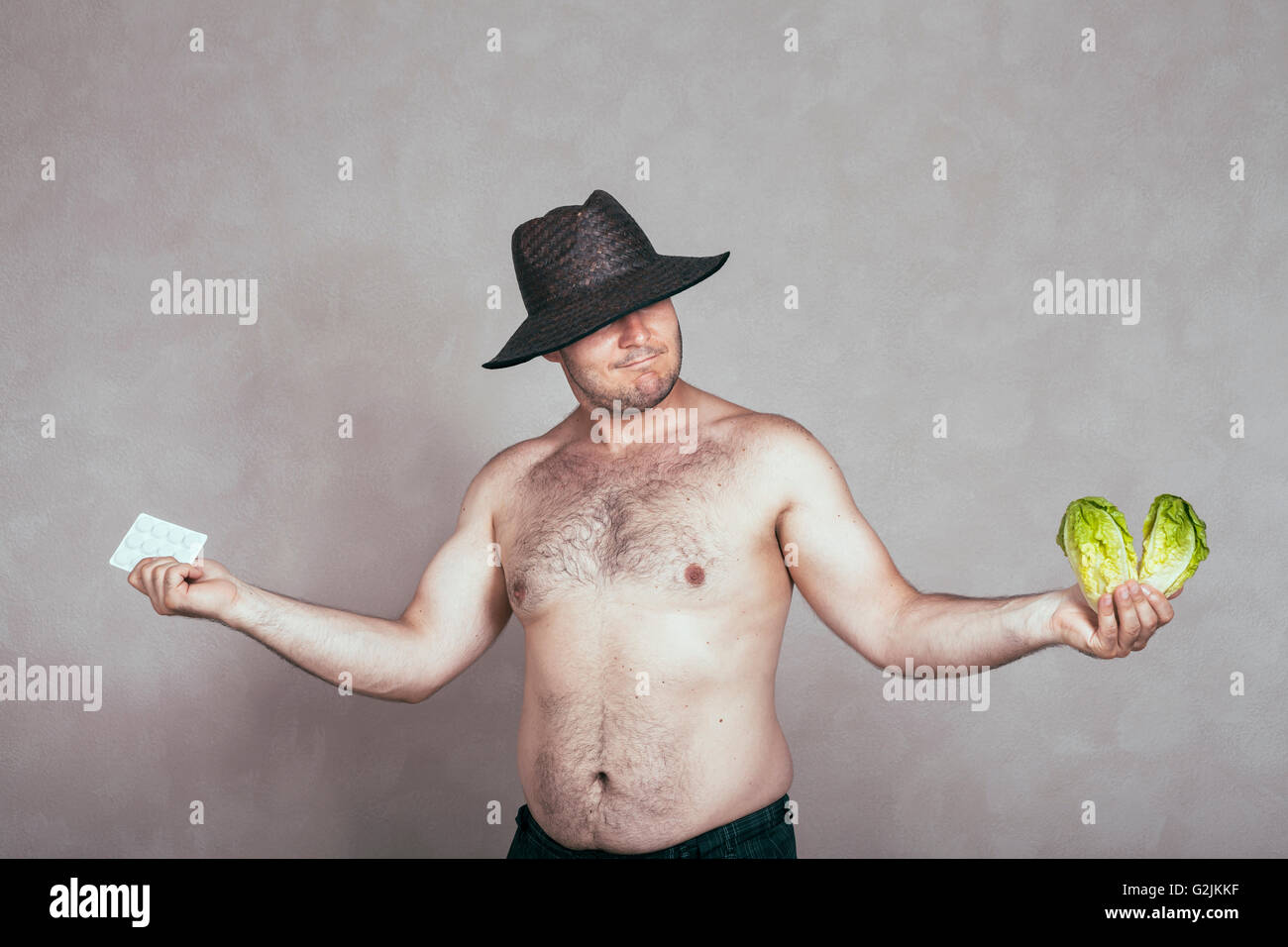 Indecisive naked corpulent man in hat holding pharmaceutical products and lettuce. Stock Photo